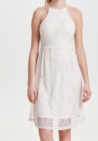 Valkoinen pitsimekko, jossa on halterneck-olkaimet, tiukka yläosa ja levenevä helma. Kankaan pinnassa on kukkakuvioita ja läpinäkyvä päällyste.