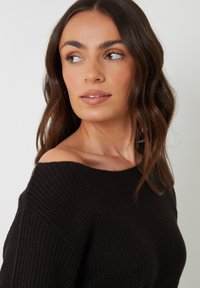 Zwarte gebreide off-shoulder trui met een losse pasvorm, voorzien van een gestructureerde stof en een brede halslijn. Zacht, chunky ontwerp.