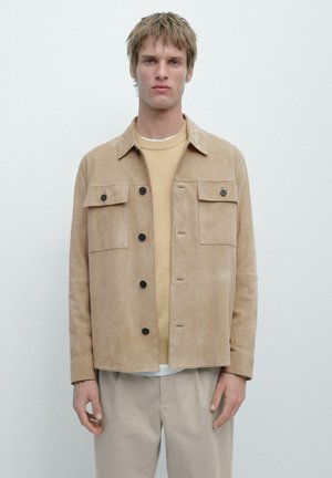 Jeune homme portant une veste en daim beige sur un pull marron clair et un pantalon beige, debout devant un fond blanc uni.
