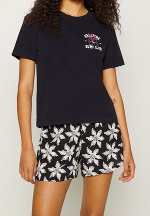 Mujer que lleva una camiseta negra "Hellfire Surf Club" y pantalones cortos negros con estampados florales blancos, de pie con los brazos relajados.
