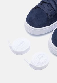 Baskets en daim marine avec semelles en caoutchouc blanches, accompagnées de lacets blancs plats placés à côté des chaussures. Le design est bas avec des orteils ronds.