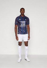 Tmavomodrý futbalový dres s vertikálnymi červenými pruhmi, vybavený golierom a logami, doplnený o biele šortky a ponožky a čierne futbalové topánky.