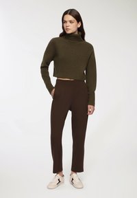 Przycięty sweter w oliwkowym kolorze z wysokim dekoltem, zestawiony z brązowymi spodniami o dopasowanym kroju. Modelka ma na sobie beżowe trampki z czarnymi akcentami.