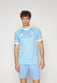Lyseblå sportsdrakt med abstrakt mønster, hvite detaljer og gule striper på skuldrene; kombinert med matchende blå shorts.