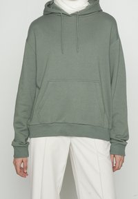 Persona con sudadera gris claro con bolsillo frontal y cordones, combinada con pantalones blancos holgados, de pie frente a un fondo liso.