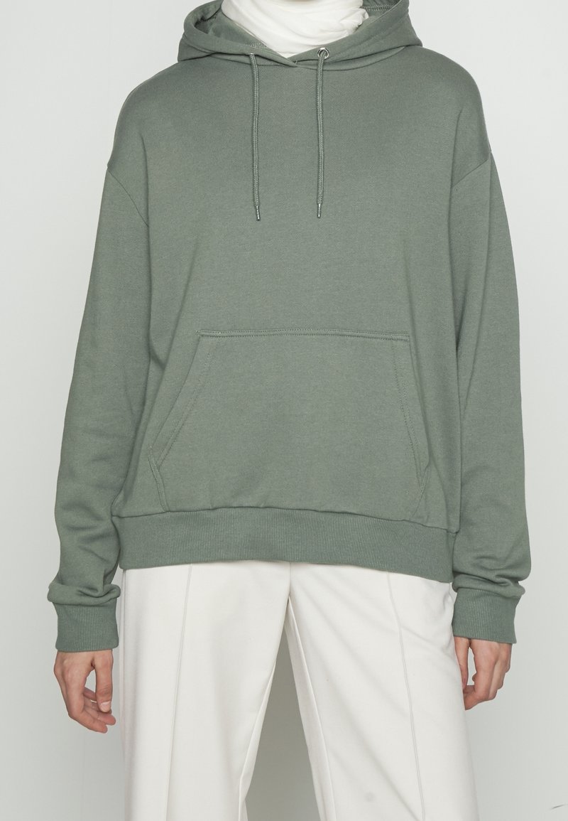 Persona con sudadera gris claro con bolsillo frontal y cordones, combinada con pantalones blancos holgados, de pie frente a un fondo liso.