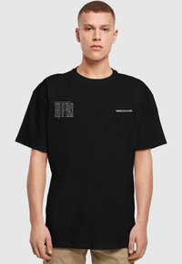 Sort bomulds T-shirt med rund hals, korte ærmer og frontgrafik: "FUGL AF FRED" til venstre og "MERCHCODE" til højre.