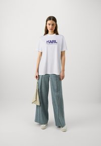 Weißes Baumwoll-T-Shirt mit lila "KARL LAGERFELD"-Aufdruck, kombiniert mit weit geschnittenen hellblauen Hosen und einer cremefarbenen Handtasche.