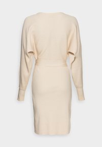 Robe beige tricotée avec une texture côtelée, manches longues et ceinture à la taille, caractérisée par une silhouette ajustée et une coupe mi-longue.