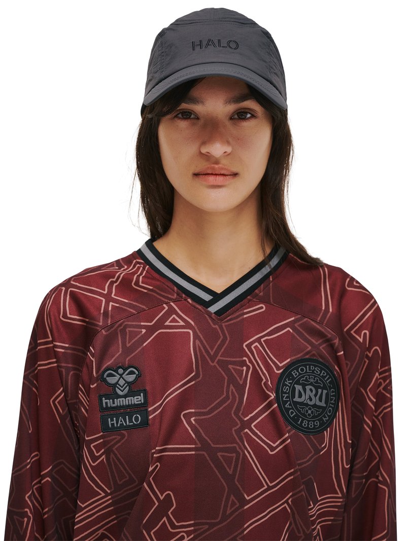 Junge Person trägt ein bordeauxfarbenes, gemustertes Sporttrikot mit dem "DBU"-Logo und eine schwarze Kappe mit dem aufgestickten Schriftzug "HALO" vor weißem Hintergrund.