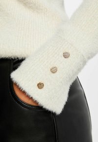 Biały, puszysty sweter o ryflowanej strukturze z trzema złotymi guzikami w kształcie serca na rękawie, w połączeniu z czarną skórzaną kieszenią.
