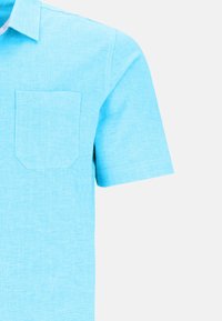 Chemise à manches courtes de couleur bleu clair, en tissu texturé, avec une poche plaquée sur le côté gauche de la poitrine et un détail de logo subtil au-dessus de la poche.