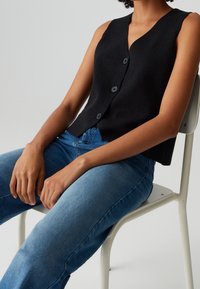 Černá bezrukávová vesta s výstřihem do V a čtyřmi knoflíkovými zapínáními, spojená s modrými denimovými džíny, sedící na židli.