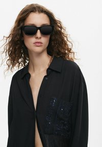 Occhiali da sole neri con una montatura rettangolare e lenti scure, abbinati a una camicia nera con bottoni caratterizzata da ornate decorazioni ricamate blu.