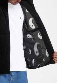 Homme en t-shirt blanc et jean bleu ouvre une veste noire montrant la doublure avec de grands symboles yin-yang blancs et noirs.