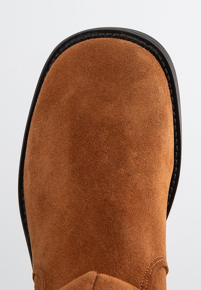 Braune Wildleder-Ankle Boots mit runder Spitze, genähtem schwarzen Sohlenrand und glatter Oberfläche, die ein minimales Design ohne sichtbare Metallteile aufweisen.