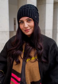 Zeszyty czarny beret, brązowo-różowy wzorzysty szalik z żółtym akcentem, nałożony na ciemny płaszcz, z falującymi, długimi brązowymi włosami.