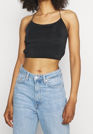 Kobieta ubrana w czarny krótki top na cienkich ramiączkach i jasnoniebieskie, z wysokim stanem dżinsy, stojąca na jednolitym tle.