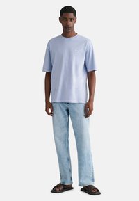Světle modré bavlněné tričko s kulatým výstřihem a krátkými rukávy, spárované se světle modrými džíny a tmavými sandály. Na hrudi je jemné logo.