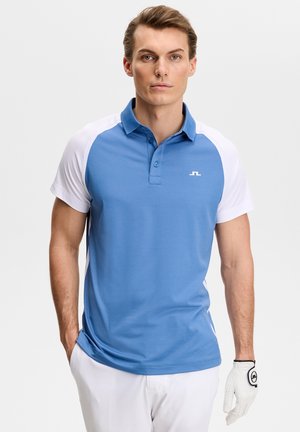 Mann trägt ein blau-weißes Polohemd und weiße Hose mit einem weißen Golfhandschuh an der linken Hand, steht vor einem einfarbigen Hintergrund.