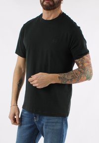 Camiseta de algodón negra con escote redondo y mangas cortas, con un pequeño logo en el pecho, combinada con jeans azules.
