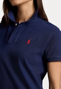 Námořnická modrá polo košile z texturované bavlny, s límcem na knoflíky a červeným vyšitým logem na hrudi. Krátké rukávy a klasický střih.