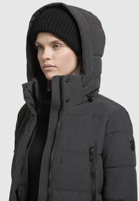 Black puffer jacket with a high collar and large hood, featuring vertical stitching, zippered side pockets, and a ribbed black beanie.