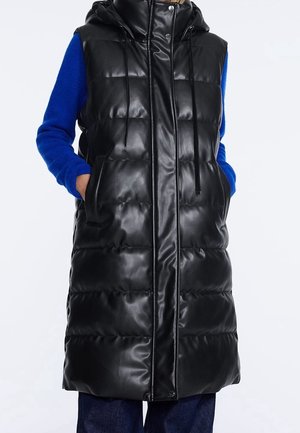 Schwarze, puffy Weste mit Kapuze, aus glänzendem Material. Mit seitlichen Taschen, Kordelzügen und langem Design, kombiniert mit einem blauen Pullover.