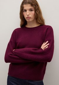Burgunderfarbener Strickpullover aus weichem Stoff, mit rundem Ausschnitt, gerippten Bündchen und einer lockeren Passform. Das Modell hat die Arme verschränkt.
