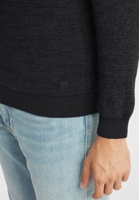 Schwarzer Pullover mit strukturiertem Strickdesign, gerippten Bündchen und Saum. Kleine Markenlabel unten links sichtbar. Tragen mit hellblauen Jeans.