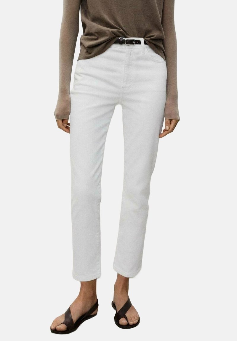 Personne portant un jean blanc à jambes droites, un haut marron à manches longues, une ceinture noire et des sandales noires, debout devant un fond uni.