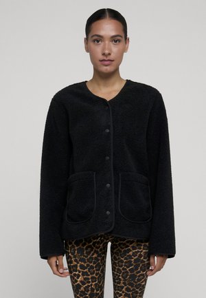 Femme aux cheveux foncés tirés en arrière, portant une veste polaire noire boutonnée et un legging imprimé léopard, face à un fond uni.