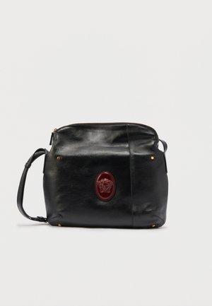 Black leather crossbody bag with adjustable strap and red oval emblem featuring a Medusa head at center front.