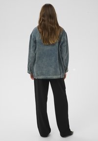 Jeansjacke in verblasstem Blau mit sichtbaren Nähdetails und lockerer Passform, getragen über schwarzen, weit geschnittenen Hosen. Strukturiertes Gewebe.