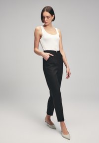 Witte geribbelde tanktop, hoge zwarte broeken met zijzakken en een gouden knop, gecombineerd met lichtgrijze puntige hakschoenen. Gladde textuur.