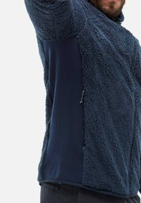 Mężczyzna w granatowej polarowej kurtce z fakturowanymi panelami i zamykaną kieszenią z boku, podnoszący jedną rękę na jednolitym tle.