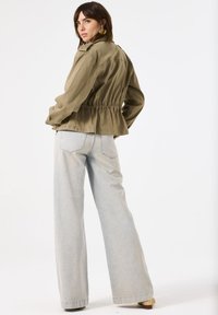 Olijfgroene cropped jacket met elastische taille, gecombineerd met lichtblauwe wijdere jeans, die een ontspannen pasvorm en zachte textuur tonen.