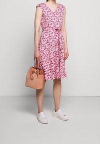 Žena v růžových květovaných šatech bez rukávů, která drží světle hnědou semišovou kabelku, ve bílých teniskách, stojící proti jednobarevnému šedému pozadí.