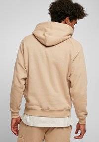 Felpa beige con cappuccio realizzata in materiale morbido, dotata di una tasca a marsupio, polsini a costine e una vestibilità rilassata, mostrata di spalle.