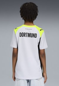 Camiseta deportiva blanca con acentos amarillos y negros, con "DORTMUND" impreso en la parte posterior. Fabricada con un tejido transpirable y texturizado.