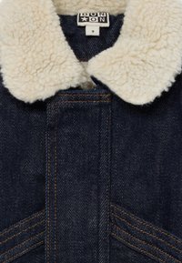 Chaqueta de mezclilla con cuello de sherpa blanco y costuras naranjas visibles en la parte delantera, mostrando la etiqueta de la marca dentro del cuello.
