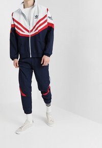 Chándal navy con franjas diagonales rojas y blancas, con chaqueta con cremallera y pantalones ajustados. Combinado con zapatillas blancas.