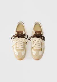 Zapatillas de ante beige y piel crema con cordones y acentos marrones, que cuentan con detalles en dorado y un interior acolchado.