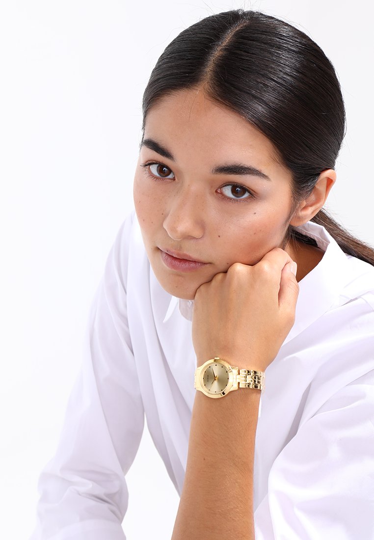 Gold metal watch with a round face and link bracelet, featuring a minimalist dial and three hands. Model wearing a white shirt.