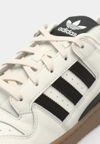 Bijele kožne tenisice s crnim antilop prugama, kremastim vezicama i crnim jezikom na kojem se nalazi Adidas logo. Guma potplat i perforirani detalji.