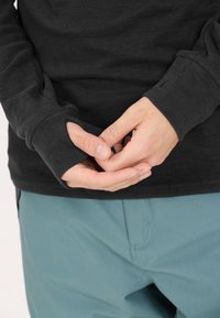 Schwarzes Langarmshirt mit Daumenlöchern an den Bündchen, kombiniert mit türkisfarbenen Hosen. Das Material wirkt glatt und enganliegend, mit minimalen Nähten.