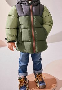 Chaqueta acolchada verde y gris con capucha, que presenta un cierre naranja y botones de presión. Combinada con jeans azules arremangados y botas resistentes.