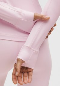 Parte superior deportiva de manga larga en color rosa, con un diseño ajustado, orificios para los pulgares en los puños y tela suave y elástica. Detalles de costura visibles a lo largo de los brazos.