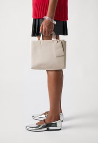 Beige läderhandväska med korta handtag, med en avtagbar etikett. Modellen bär en röd stickad tröja och en veckad grå kjol, silverfärgade skor.