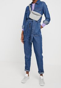 Blå denimoverall med knytbälte, i kombination med en grå axelväska och grå sneakers. Långa ärmar med uppvikta mudd och ribbade lila detaljer.